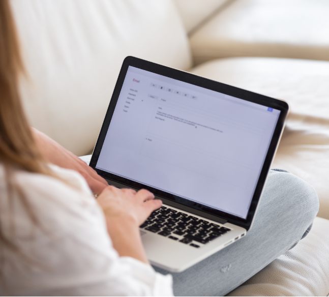 Person typing on a laptop while seated on a sofa, with a light background.