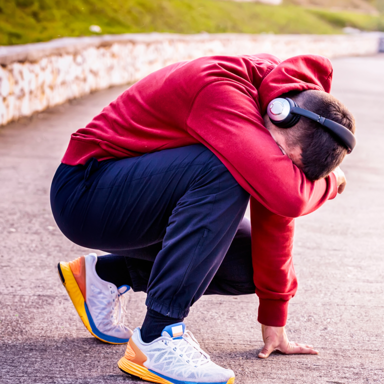 A person in a red hoodie squats down, resting with their head on their knees, wearing headphones.