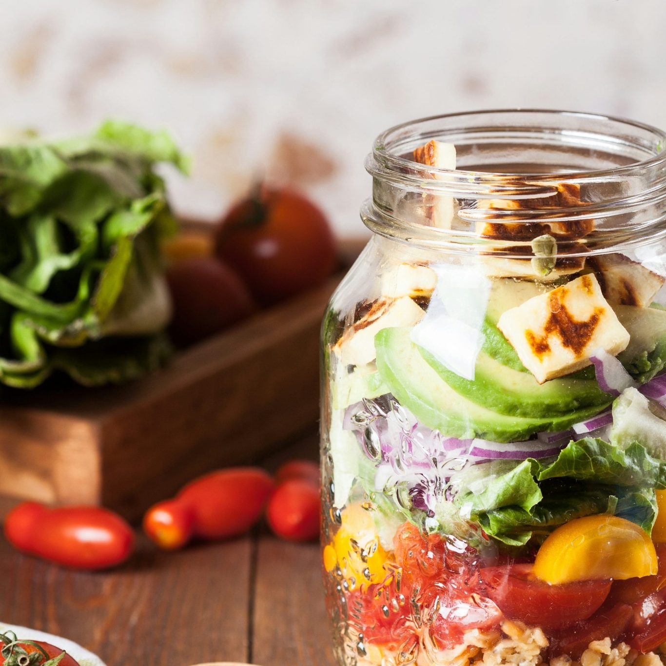 A layered salad in a jar with greens, tomatoes, cheese, and colourful vegetables.