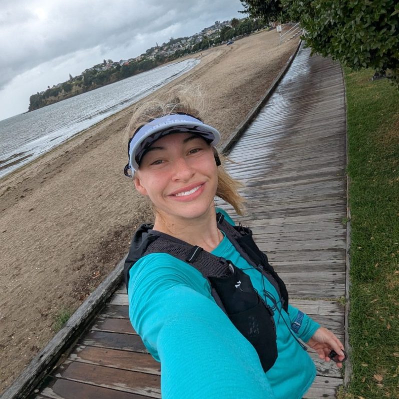 Smiling woman in a blue top and visor near a sandy beach and ocean.